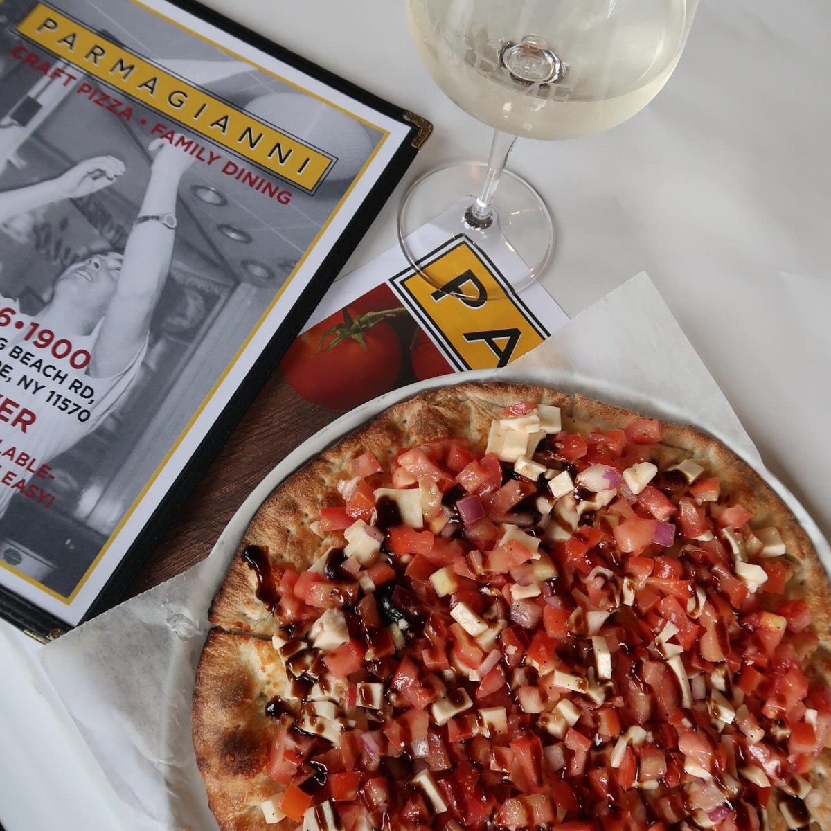 A Parmagianni craft pizza topped with diced tomatoes, fresh mozzarella, red onion, and a balsamic glaze, beside the restaurant's menu and a glass of white wine.