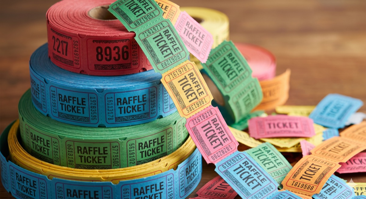 A colorful stack of red, blue, green, and yellow paper raffle ticket rolls printed with 'RAFFLE TICKET' and serial numbers, with loose tickets fanned out beside them on a wooden table.
